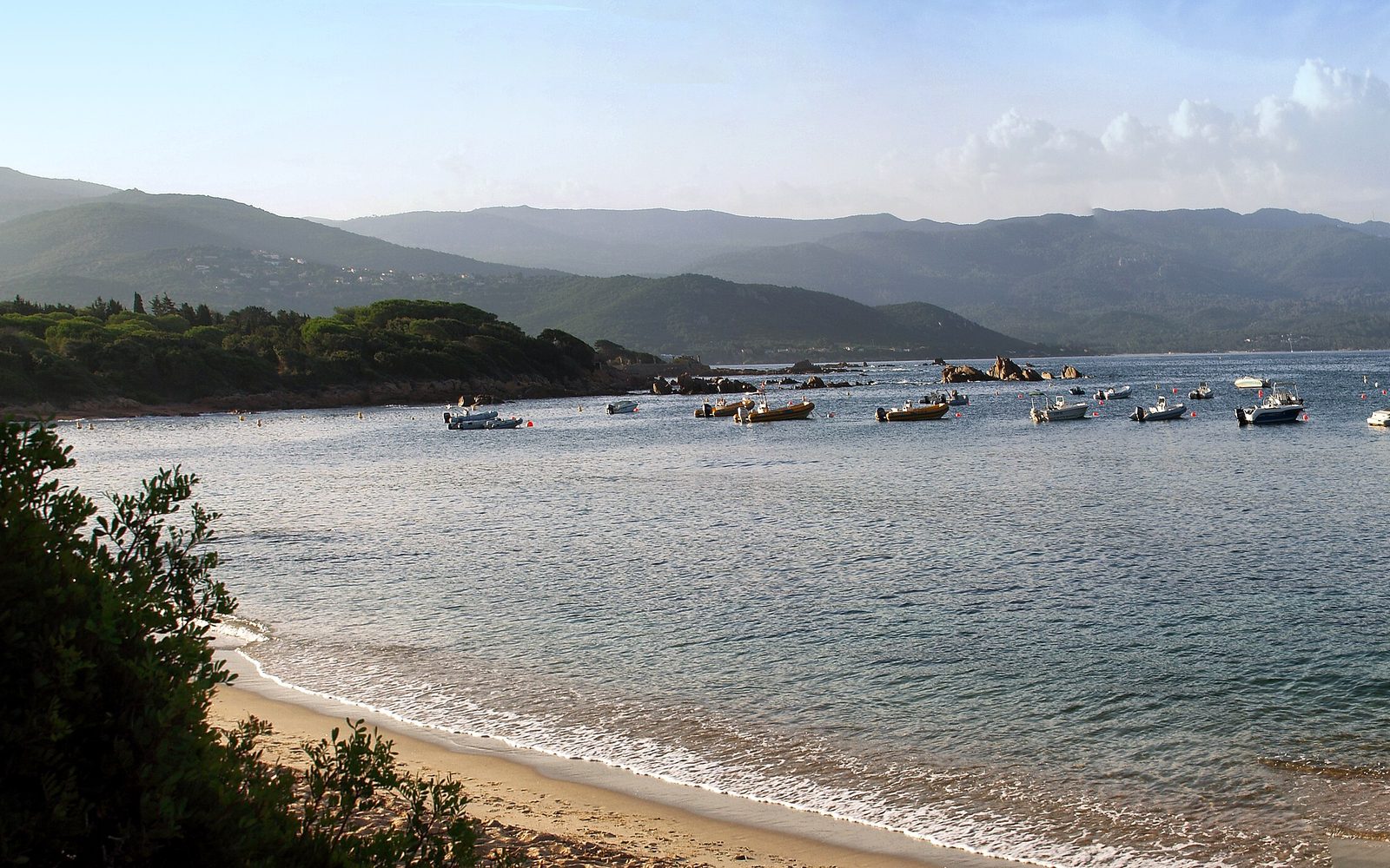 Une petite plage de la presqu’île de l’Isolella, des bateaux au mouillage et les montagnes au fond